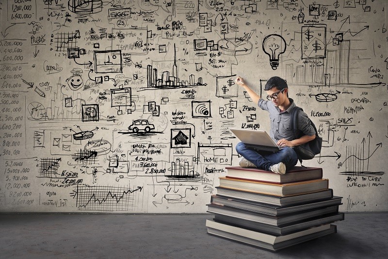 Young boy sitting on some books drawing his ideas on a wall while working on its computer